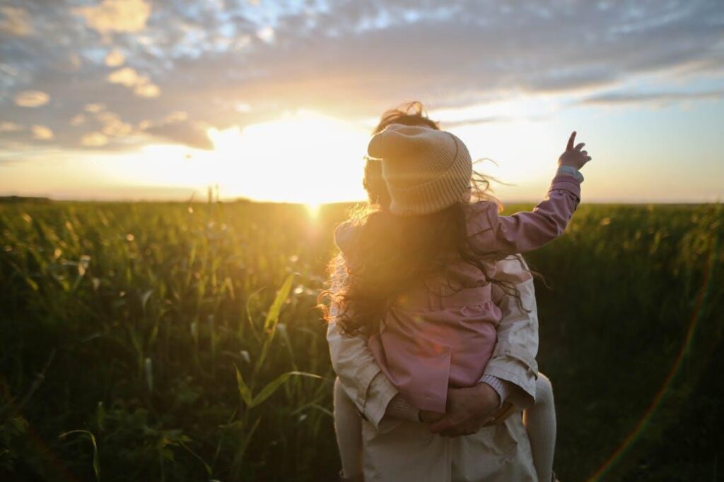 Free A mother and daughter embrace and point at the sunset in a grassy field. Stock Photo
