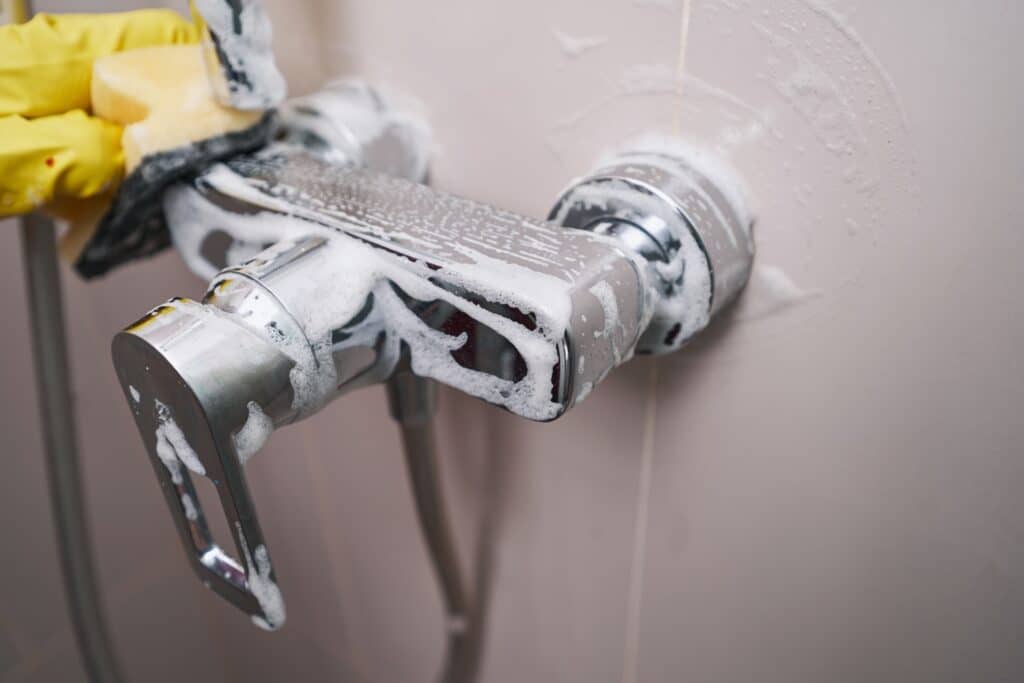 Cropped photo of gloved hand wiping bathroom tap using damp sponge and detergent